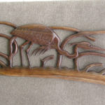 A carved wooden piece by Gilbert Carey depicting a crane crouching behind reeds.