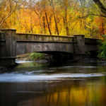 Photograph by Christopher Costa of a bridge over rippling water during autumn, with many orange and yellow trees in the background.