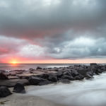 Photograph by Christopher Costa of a jetty under an orange and pink sunrise. The sky is filled with billowy clouds and fog runs over the sand.