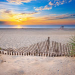 Photograph by Christopher Costa of a beach with a tan sand fence and green plants in the foreground. The blue ocean and lifeguard sit in the distance.