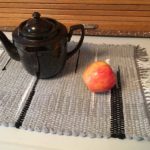 A teapot and apple sit on top of a grey handwoven placemat by Belinda DeCicco.