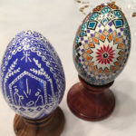 Two pysanky by Marino Diaz. The left egg has a blue nativity scene with pattern embellishments surrounding the scene. The right is decorated with a red flower and concentric multicolor shapes.