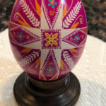 A fuchsia pysanky by Marino Diaz. The egg is decorated with a cross in the center forming a rose symbol. Images of grain and blue flowers decorate the space around it.