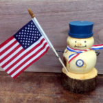 A snowman carved from a log with a navy badge and blue hat, holding an american flag. Created by Thomas Ferrie.