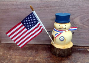 A snowman carved from a log with a navy badge and blue hat, holding an american flag. Created by Thomas Ferrie.