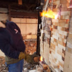 Photo of the kiln at WheatonArts, covered with bricks. A small fire spouts out around the gaps.