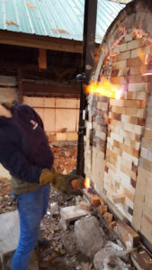 Photo of the kiln at WheatonArts, covered with bricks. A small fire spouts out around the gaps.