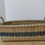 A long woven basket by Mary May with a large deep blue stripe woven around near the bottom.