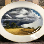 A painted porcelain plate with a scene of a storm cloud looming over the plains. Created by Michelle Post.