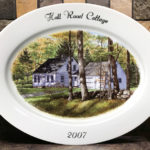 A painted porcelain plate with a scene of a home with a barn beside it, slightly obscured by trees in the large forest around it. Text at the top reads Hall Road Cottage, bottom text reading 2007. Created by Michelle Post.