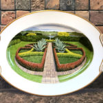 A painted porcelain plate with a scene of a well manicured garden with tall hedges and leafy green plants, a peek of a lake in the distance. Created by Michelle Post.