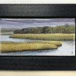 A painted piece of porcelain in a thick black frame showcasing a scene of a river winding through the marsh. Created by Michelle Post.