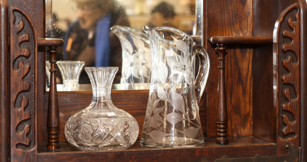 Two clear glass vessels sitting on an antique wooden shelf with a mirror behind them.