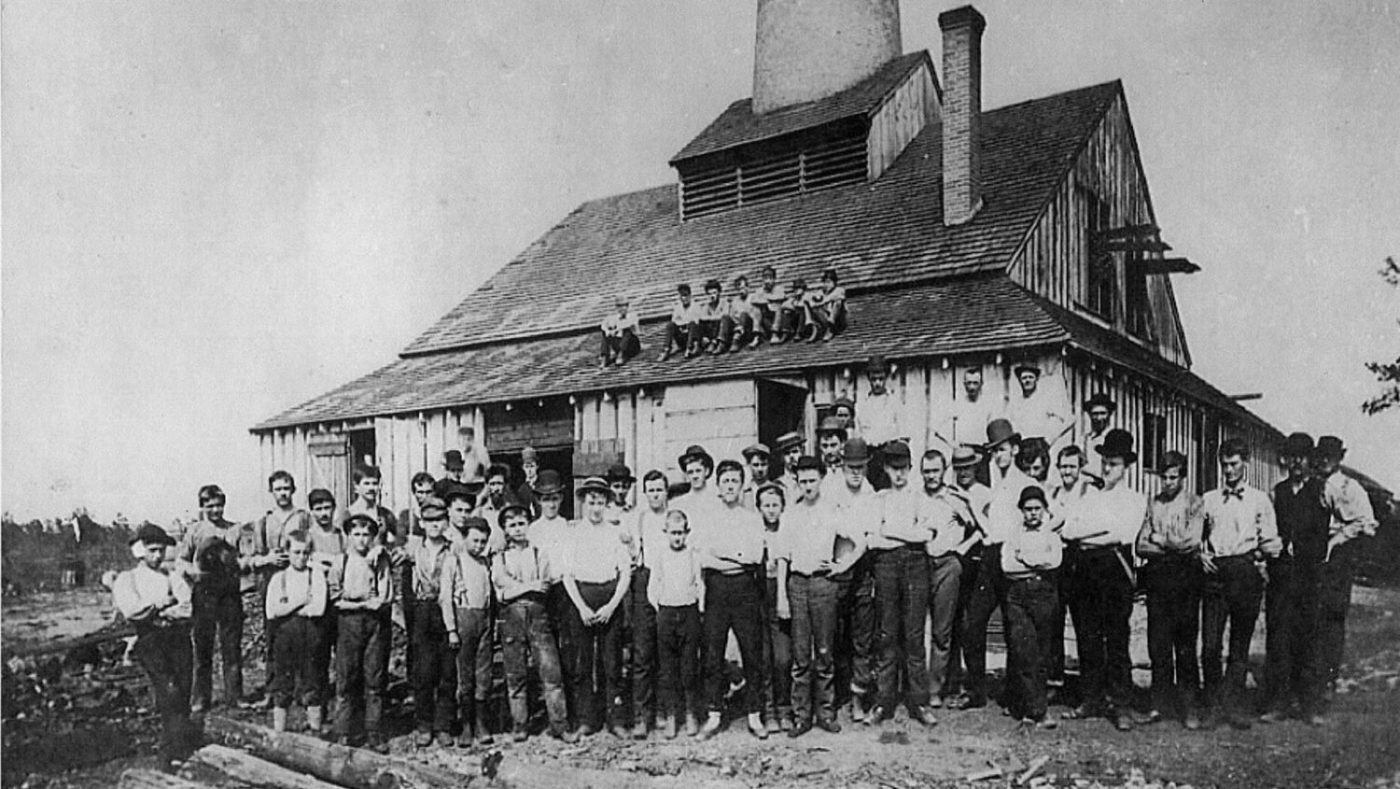 A photo of T.C. Wheaton Glass Company employees in 1888, gathered in front of the factory. A few employees sit on the roof. They all wear white shirts and dark pants.