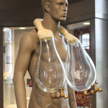 Allan Wexler, Vessicles. Around a mannequins neck is a large vessel with two glass containers on either side. At the end of each is a bronze faucet.