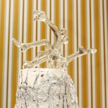 A bundle of flailing limbs come out from a central point, created in clear glass. The piece sits on a white piece of wood.