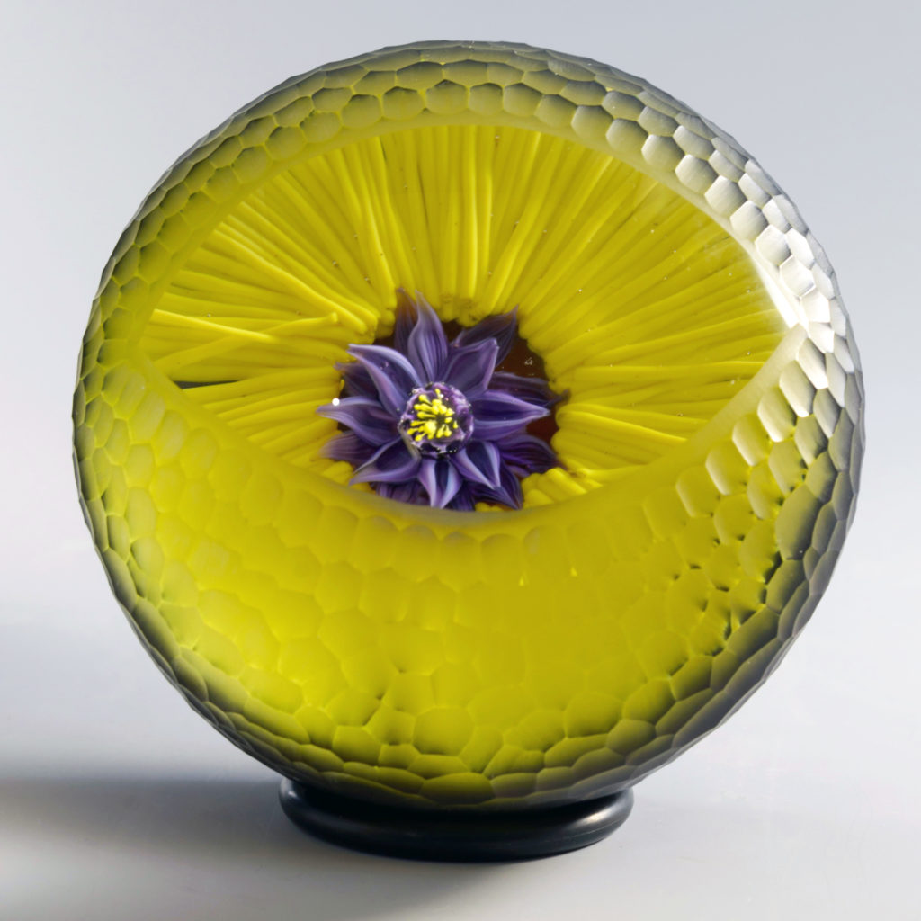A paperweight by Melissa Ayotte, entitled 'Watchful Eye', with a purple flower in a bright yellow background.