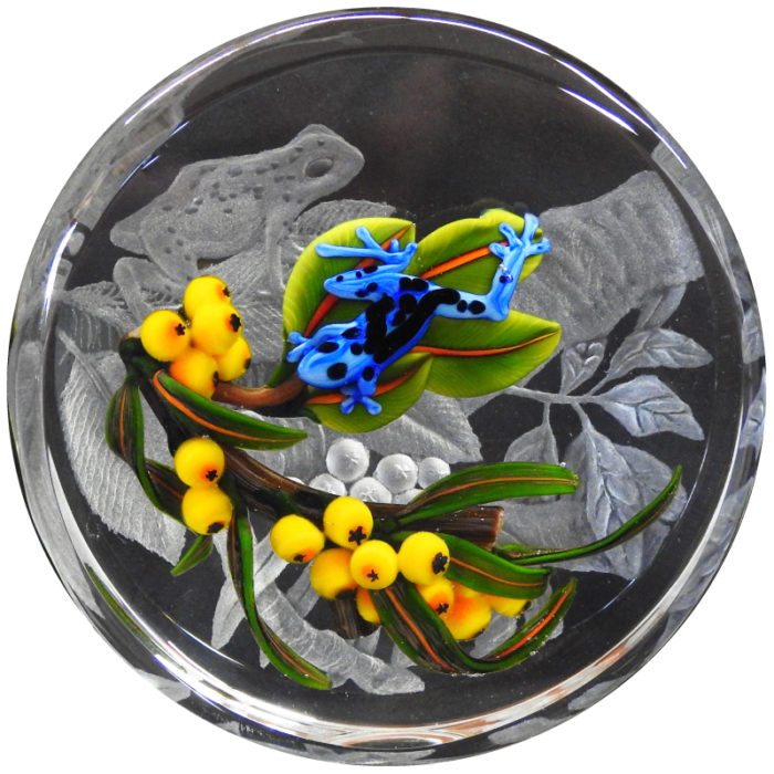 An engraved paperweight created as a collaboration between Cathy Richardson and Colin Richardson. On top of a grey engraved depiction of a frog on a branch, there is a colorful blue frog on a similar branch with bright yellow berries.