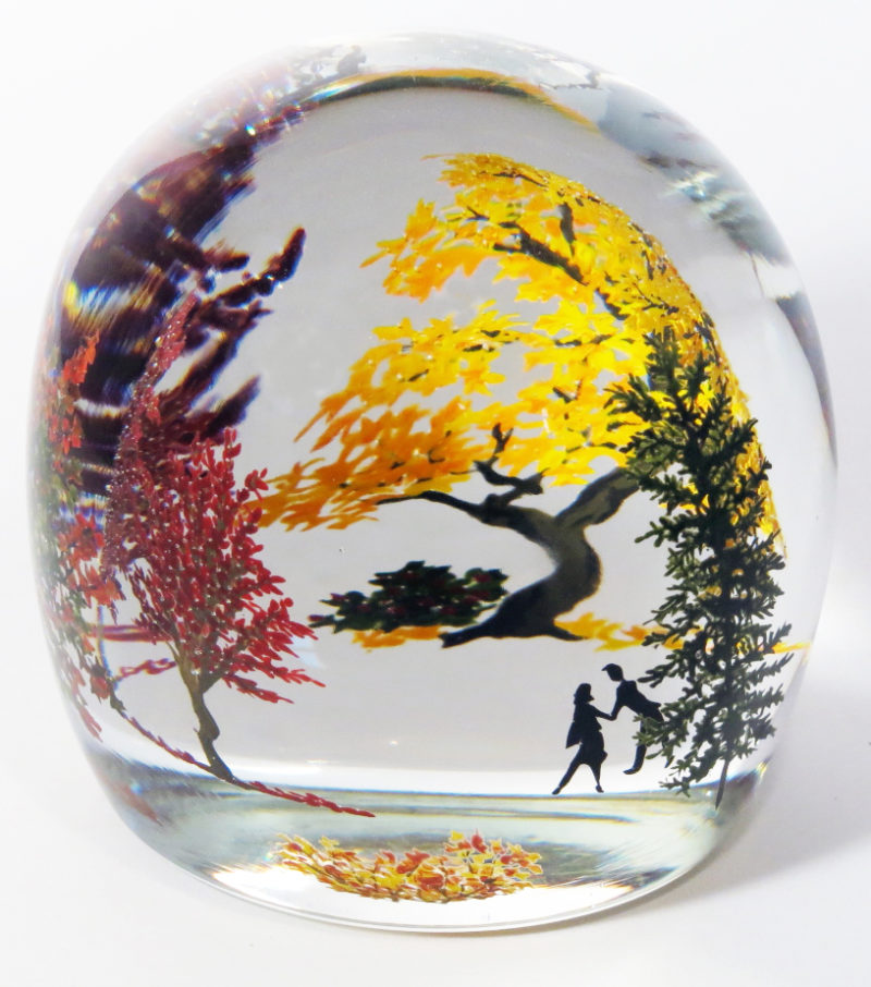 A paperweight by Alison Ruzsa. The tall dome is painted with a scene of a silhouetted couple walking under orange and red trees with a pile of fallen leaves in the center.