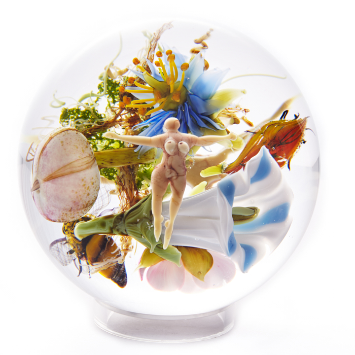 A paperweight by Paul Stankard with a floating figure in front of a bundle of flowers in white, blue, pink, and orange.