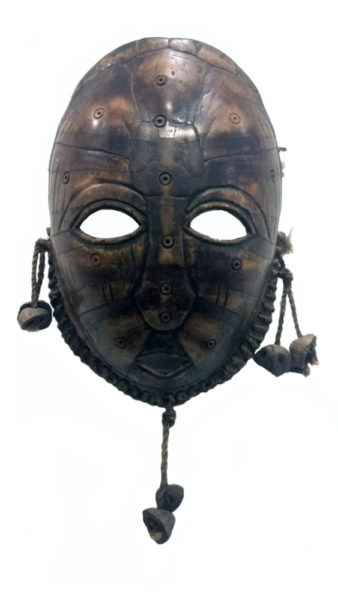 “Face” Lega Mask, Congo, artist unknown, Bwami society. Carved turtle shell, rope, seeds. Collection of Frederick Kramer. The mask is a dark black and brown with eye holes. Stones dangle from the ear and chin areas.