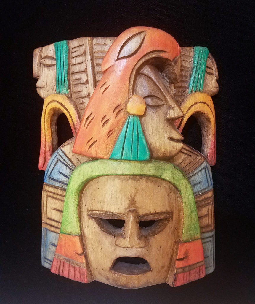 A carved, painted, wooden Mexican Mask titled "Ancestors". The mask is multicolored. Around the face of the mask are three heads with closed eyes, wearing headdresses.