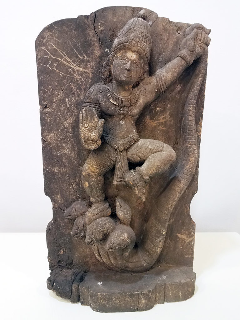 A woodcarving of Krishna, Avatar of Vishnu. Krishna has a hand out forward and the other up and out, holding up a snake.