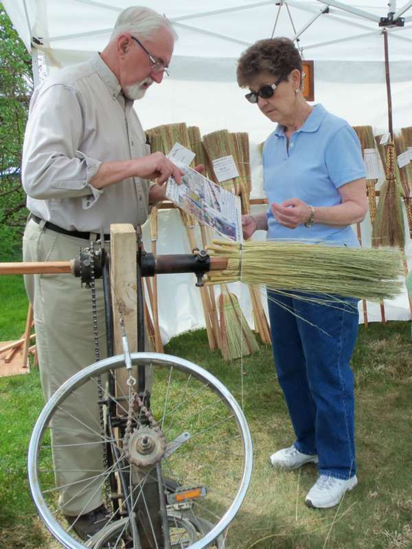 Dr. Sam Moyer with his"broom winder" explaining the process with a visitor, surrounded by brooms.