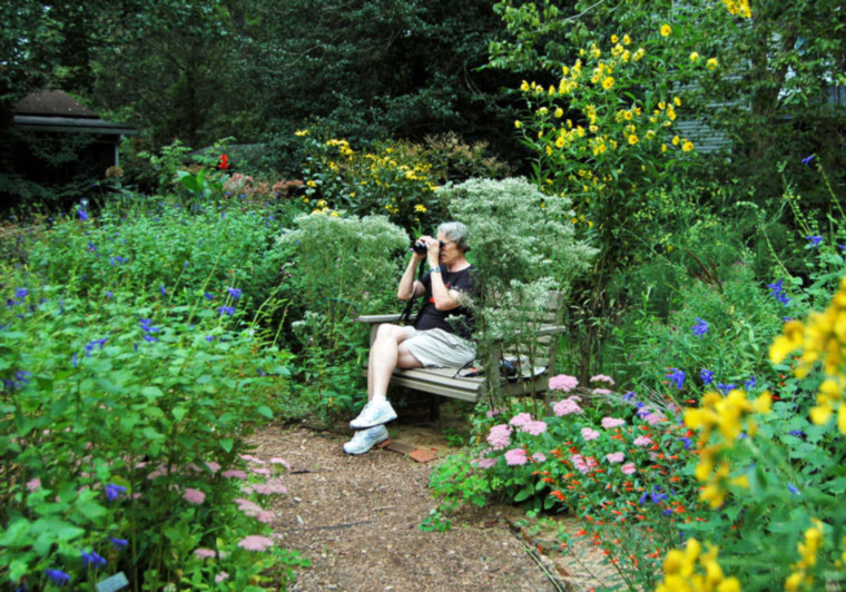 Pat Sutton sitting on a bench looking through binoculars surrounded by flowers in her garden.