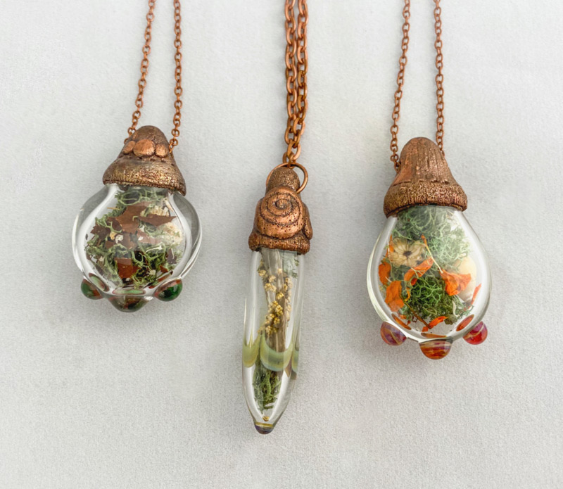 Three pendants by Dominique Jean Bebak, fashioned like terrariums with small green plants inside. Each pendant has a deep copper color cap, which holds it to the copper colored chain.
