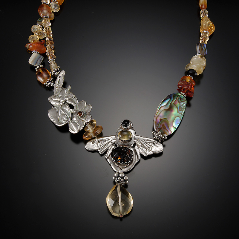 A necklace by artist Diana Contine. The necklace is full of earth toned beads of many shapes and sizes. The pendant in the center is a silver honeybee with an amber colored charm hanging beneath.