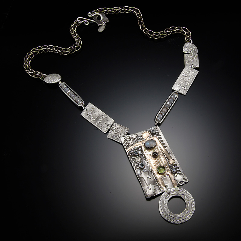 A necklace by artist Diana Contine. The necklace has a metal chain and a large rectangle pendant with small colorful stones, stamped with small lines and stars. A flat ring hangs beneath the pendant.