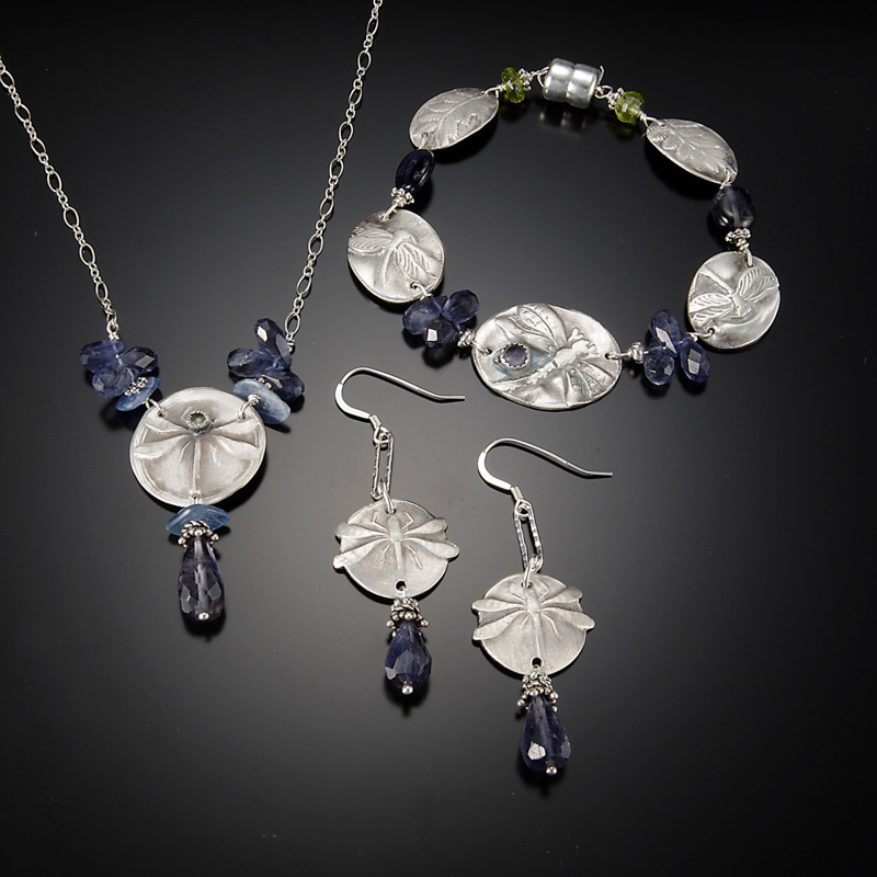 A set of earrings, bracelet, and necklace by jewelry artist Diana Contine. The necklace has a dragonfly design stamped onto a round silver pendant. Purple beads dangle below the pendant and on the silver chain. The earrings are similarly designed with a stamped dragonfly on a small circle disk and purple beads dangle below. The bracelet has purple beads connecting each of the five ovals with the dragonfly design.