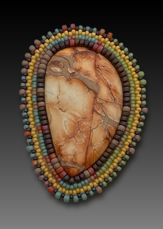 A tan and orange stone is surrounded by earthy colored beads in brown, yellow, red, and a pastel blue. Artist: Sheila Fernekes.
