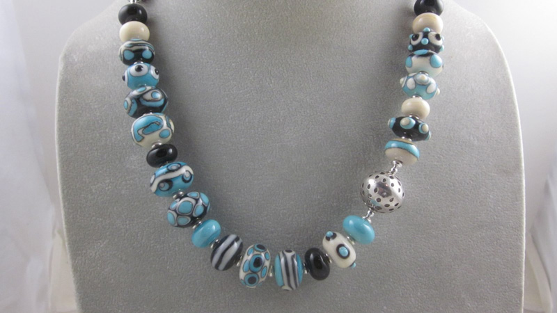 A necklace by artist Pamela Iobst. Teal, white, and black circular flamework beads drape around the neck of the display.