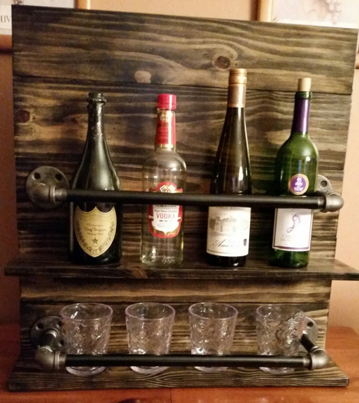 A brown wooden wine rack by Sharon Knapp holds four bottles on the top row and small glasses beneath.