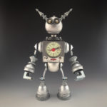 A metal found object sculpture by Brian Marshall. The sculpture resembles a robot with a timer in its chest. Its body is made of miscellaneous metal pieces.