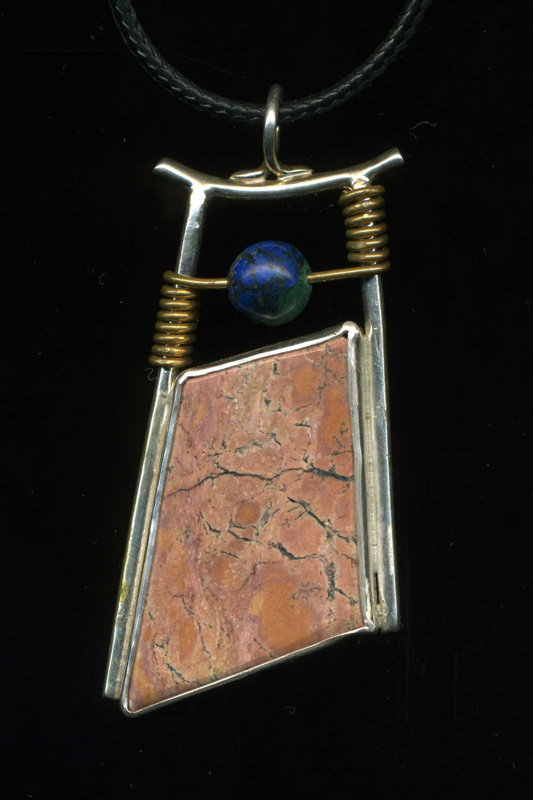 A pendant by artist Anthony Niglio. A rectangular tan stone has a silver border around it. A small blue bead sits above the stone, connected to the pendant with a copper color wire.