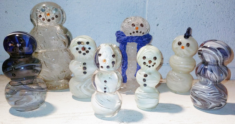 8 little glass snowmen by Jarryd Pezzillo. Some snowmen are in a milky, swirling white. The two on each end are in a swirled white, black, and blue. The snowman in the center wears a blue scarf and a deep frown.