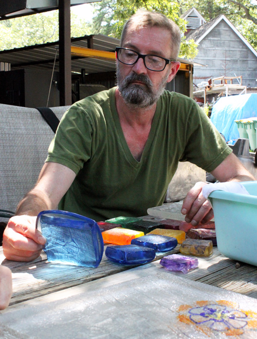 Artist Virgil Marti looks at a piece of translucent blue glass that he is holding. In front of him are several pieces of thick glass in different colors.