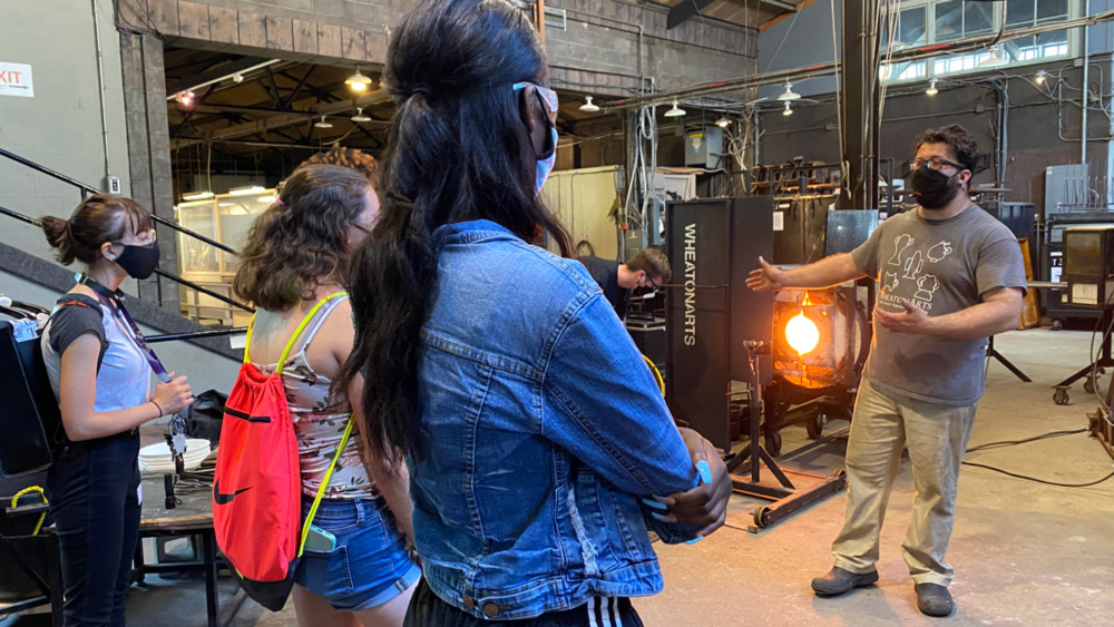 Rowan University students listen to Glass Studio Artist Skitch Manion, speaking in the WheatonArts Glass Studio.