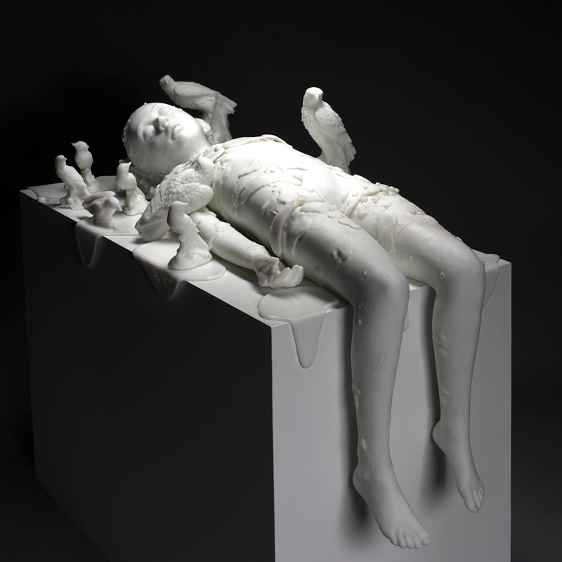 "Snow Child III", a cast glass sculpture by artist Sibylle Peretti. The pure white sculpture depicts a child laying back, legs dangling over the white surface. The figure is surrounded with similarly sculpted white birds.