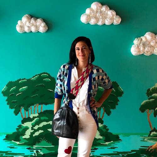 Artist Erica Rosenfeld stands in front of a colorful green backdrop, depicting clouds and painted trees.