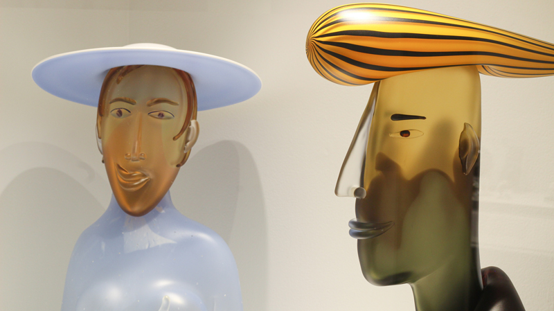 Detail of two blown glass sculptures by Artist Dan Dailey. "Curious" (left) is a glass sculpture of a person smirking, wearing a baby blue outfit and sun hat. To the right is a profile view of "Forward", depicting a person with a black shirt and orange and black striped hair.