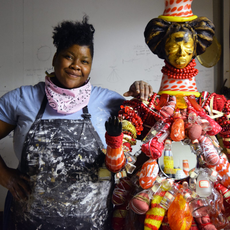 Vanessa German stands next to a large sculpture wearing bright red clothing made of found objects.
