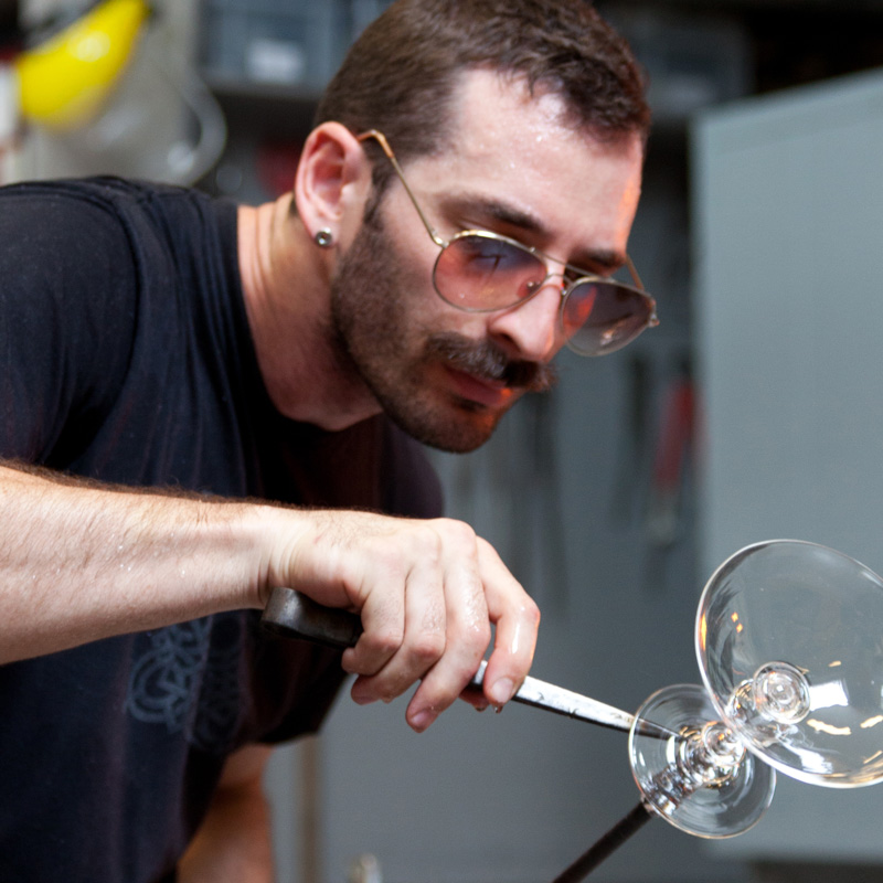 Closeup of Glass Artist Alexander Rosenberg carefully shaping clear glass on the end of a blowpipe.