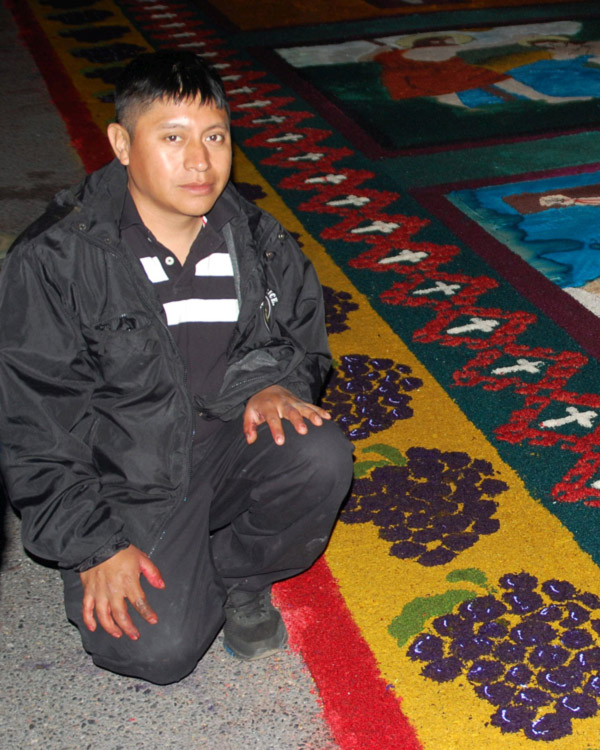 Artist Ubaldo Sanchez kneels in front of a colorful sawdust carpet.