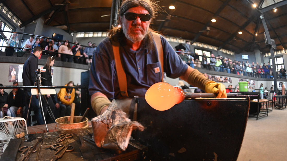 Glass artist Don Friel sitting at the Glass Studio bench with wet newspaper in one hand as he rotates hot glass on the end of a blow pipe in front of a crowd.