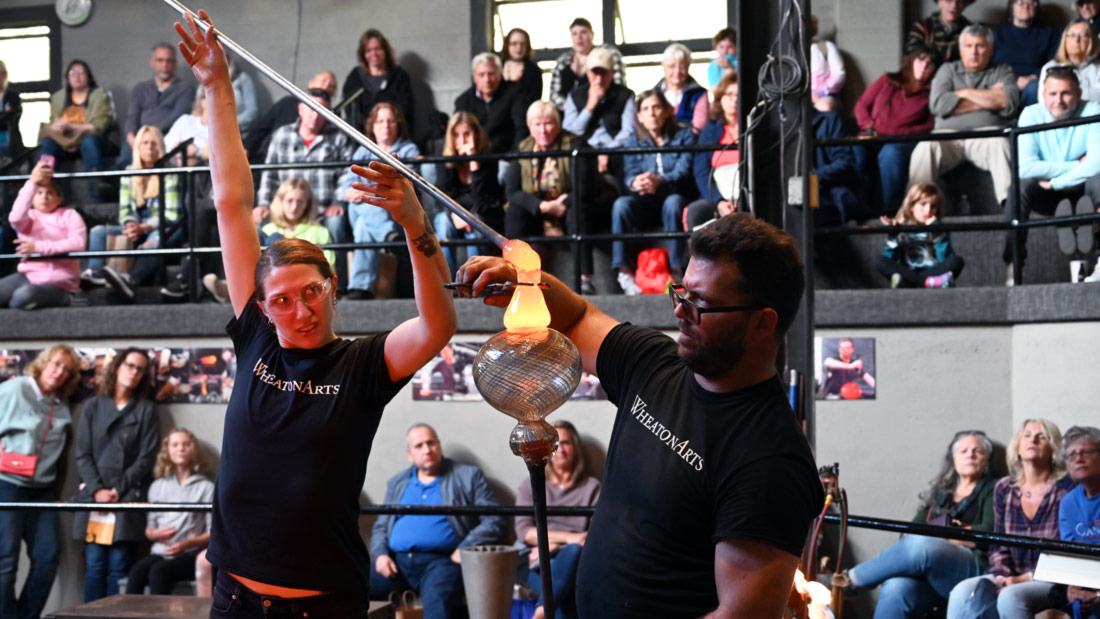 Two Glass Studio Artists use molten glass to shape a vessel in front of a crowd of spectators.