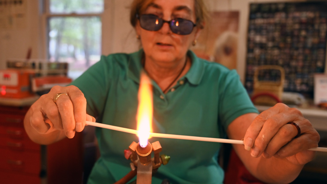 Flameworking Artist Susan Boyce carefully holds a cane of glass over the flame of a torch.